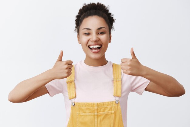 Portrait of attractive african woman in trendy yellow overalls, showing thumbs up