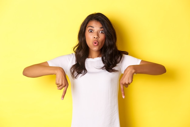 Portrait of attractive africanamerican girl pointing fingers down and looking amazed