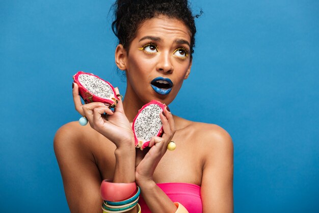 Portrait of attractive afro american woman with fashion makeup holding ripe pitaya cut in half in both hands isolated, over blue wall
