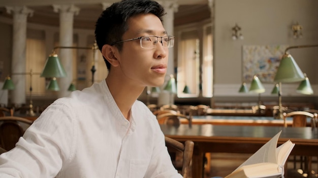 도서관에서 꿈꾸는 듯한 시선으로 교과서를 들고 있는 매력적인 아시아 학생 초상화 대학에서 공부하는 청년