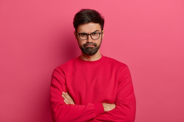 Portrait of attractive bearded young man keeps hands crossed, makes negative opinion about something, smirks from dissatisfaction, annoyed someone tells lies, wears glaasses and red sweater.