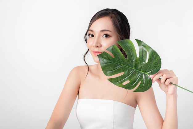 化粧品や健康メディアの白い背景に白いドレスを着て有機緑の葉でポーズをとってファッションの魅力的な美しさのアジアの女性の肖像画