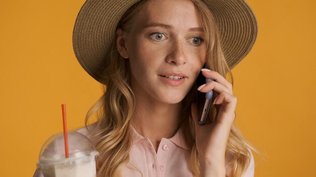 Портрет привлекательной блондинки в шляпе, разговаривающей по мобильному телефону на желтом фоне