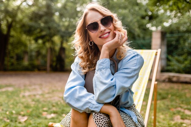 Портрет привлекательной белокурой улыбающейся женщины, сидящей на шезлонге в летнем наряде