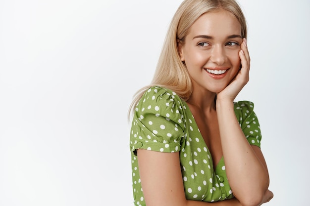 Free photo portrait of attractive blond woman in dress touching face and smiling coquettish standing over white background