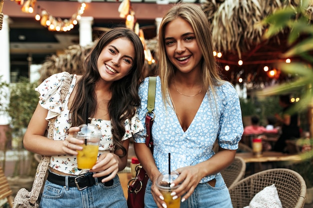 Portrait of attractive blonde and brunette girls in stylish floral blouses and denim pants smiling widely and holding lemonade glasses outside