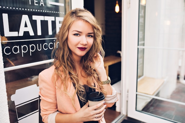 Portrait attractive blonde girl with long hair chilling on terrace in cafe. She has vinous lips, holds cup