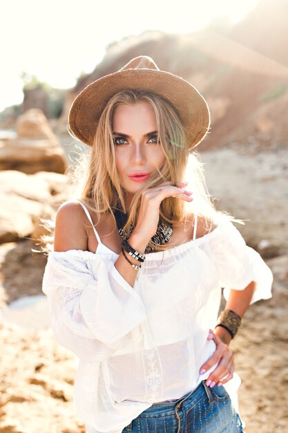 Portrait of attractive blonde girl with long hair in hat posing to the camera on beach. She is looking to the camera.