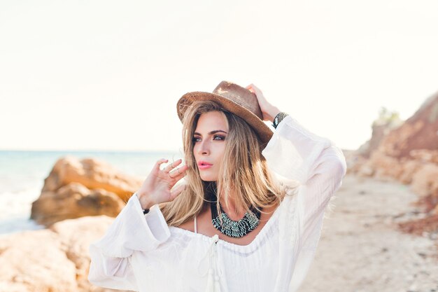 Portrait of attractive blonde girl with long hair posing to the camera on rocky beach. She wears white shirt, hat, ornamentation.