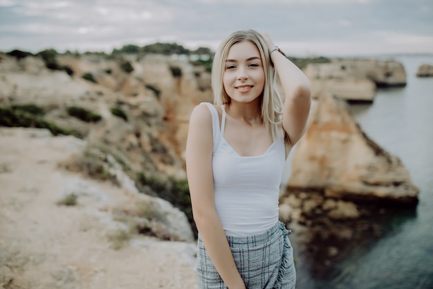 Free photo portrait of attractive blonde woman posing wears straw hat on rocky beach on sunset