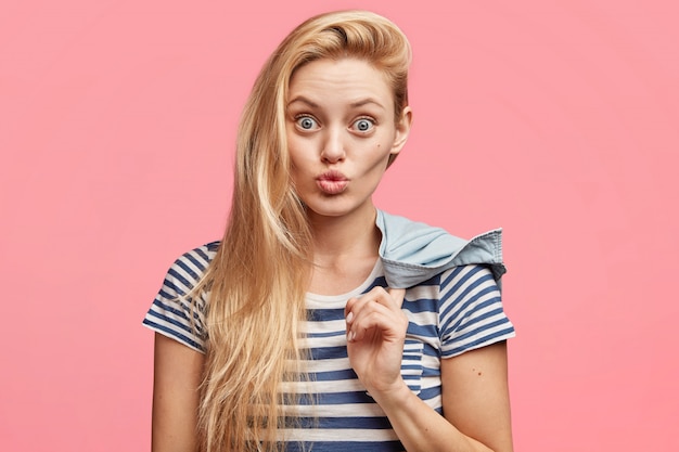 Portrait of attractive blonde young woman with pleasant appearance, wears striped t shirt, has round lips, stares at camera, makes grimace