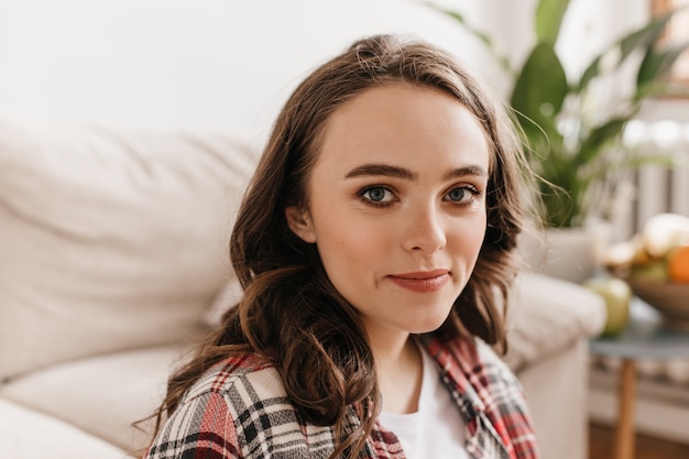 Portrait of attractive blue-eyed brunette curly woman in red plaid shirt smiling and looking into front in cozy light living room