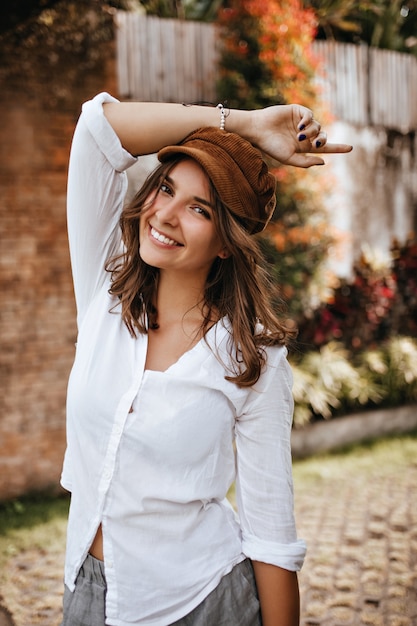 Portrait of attractive brown-eyed young woman in oversized cotton shirt and corduroy hat with smile looking at camera in yard.