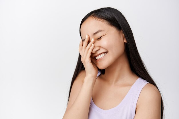 Portrait of attractive brunette asian girl, laughing and smiling happy, holding hand on face, standing in tank top on white