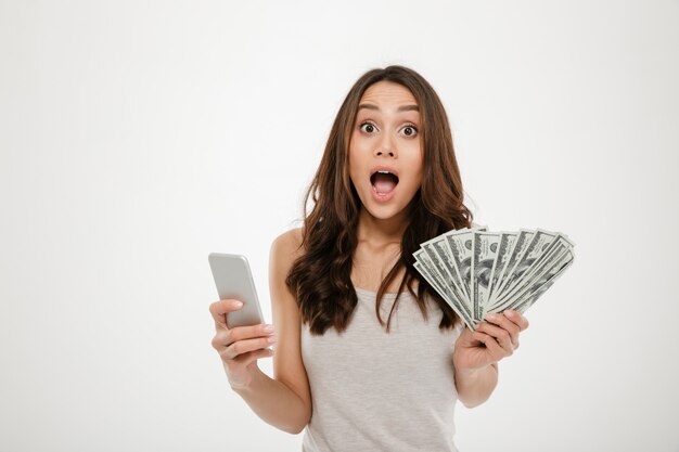 Portrait of attractive brunette female 30s winning lots of money dollar currency using her smartphone, being joyful over white