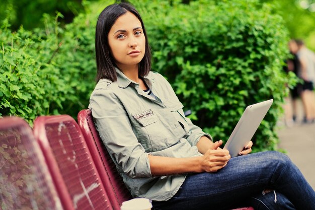 매력적인 갈색 머리 여성의 초상화는 벤치에 앉아 여름 공원에서 태블릿 PC를 보유하고 있습니다.