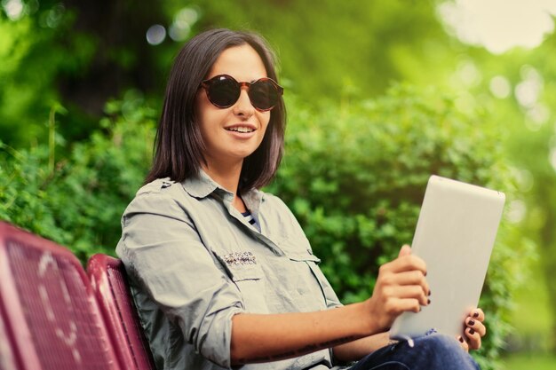 Portrait of attractive brunette female sits on a bench and holds tablet PC in a summer park.