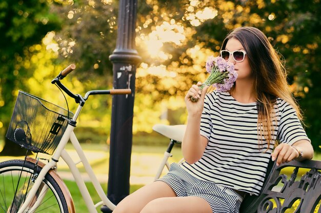 Portrait of an attractive brunette sits on a bench with a bicycle in a city park.