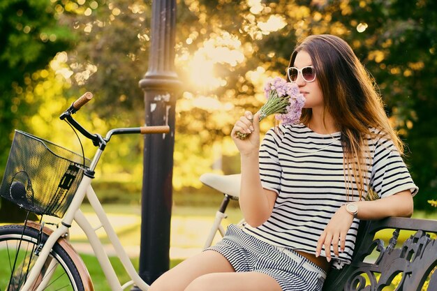 Portrait of an attractive brunette sits on a bench with a bicycle in a city park.