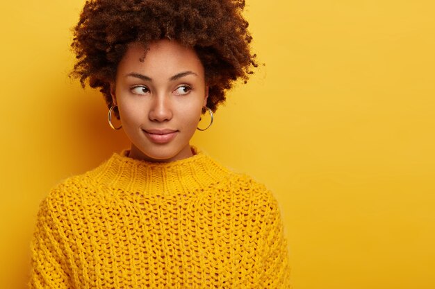 Portrait of attractive brunette woman peeks aside, has serious face expression, stands alluring against yellow background, wears no makeup, dressed in warm sweater