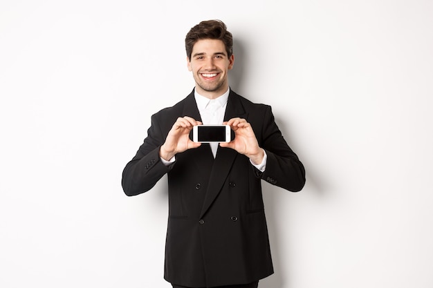 Portrait of attractive businessman in black suit, holding smartphone horizontally and showing screen, smiling pleased, standing against white background