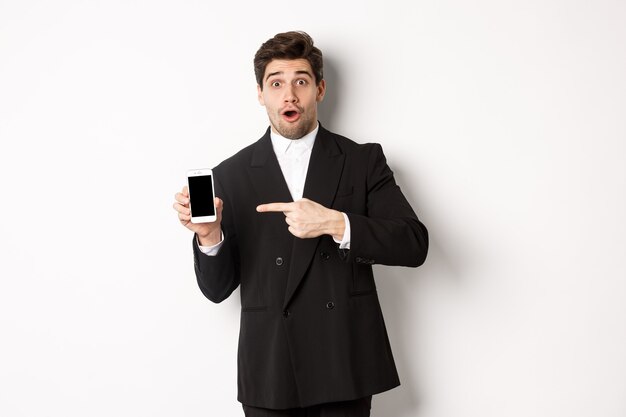 Портрет привлекательного бизнесмена в черном костюме, удивленного и указывающего пальцем на экран смартфона, стоящего на белом фоне