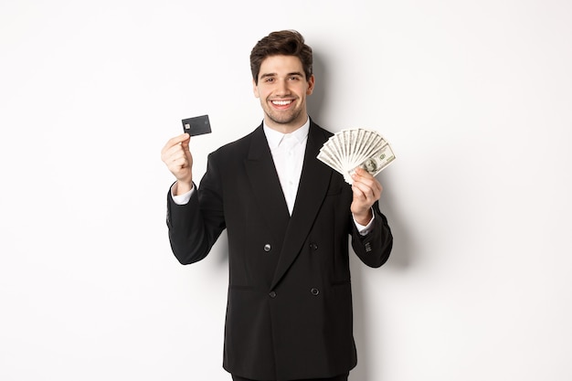 Portrait of attractive businessman in black suit, showing money and credit card, smiling pleased, standing against white background