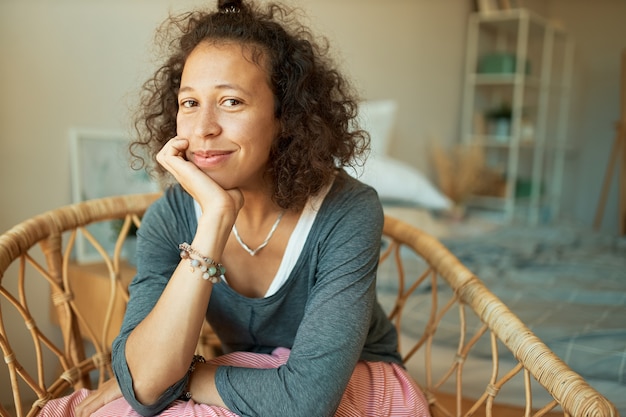 Free photo portrait of attractive casually dressed young woman with wavy hair and no make up sitting on basket armchair