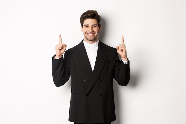 Portrait of attractive caucasian man in stylish black suit, pointing fingers up and smiling, showing christmas advertisement, standing over white background.