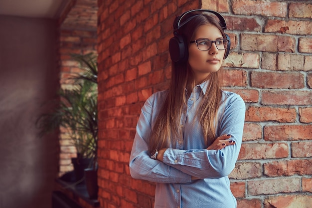 Portrait of an attractive charming brunette in glasses and blue shirt crossed arms and listening to music on headphones leaning against on a brick wall in a room with loft design.