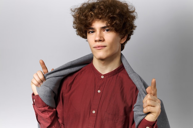 Portrait of attractive cool male student wearing shirt and gray jacket on shoulders raising fore fingers, pointing up, showing upwards, staring at camera with confident look, posing isolated