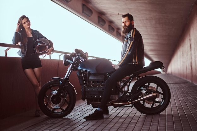 Portrait of an attractive couple - brutal bearded biker in black leather jacket sitting on a motorcycle and his young sensual brunette girlfriend on a footway under a bridge, looking at a camera.