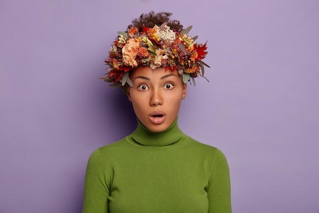 Portrait of attractive curly haired adult woman looks with shock at camera, feels stupefied and amazed, keeps mouth opened, wears autumn wreath on head, poses in studio against purple background