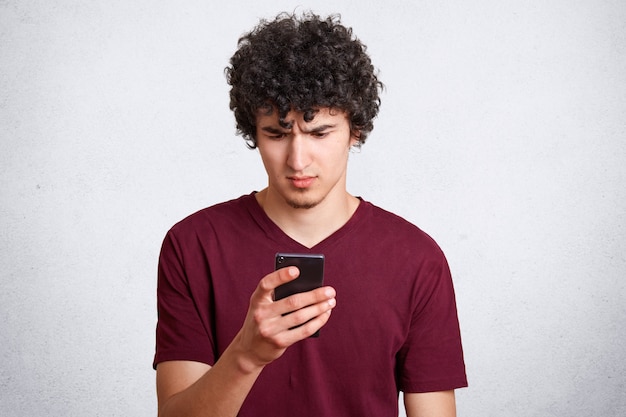 Portrait of attractive curly male looks in bewilderment on mobile phone