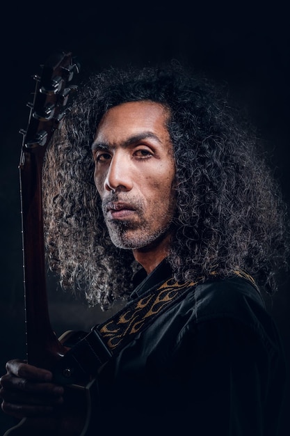 Portrait of attractive curly man with guitar at dark photo studio.