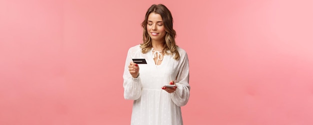 Portrait of attractive and cute blond girl with short curly hairstyle white dress holding smartphone and credit card insert digit numbers to register in shopping app buy online pink background