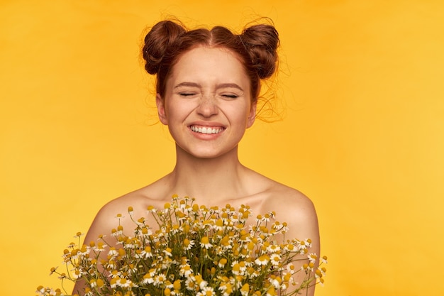 Portrait of attractive, cute, charming, red hair girl with buns. holding a bouquet of wildflowers and squints in a smile. healthy skin. closeup, stand isolated over yellow wall