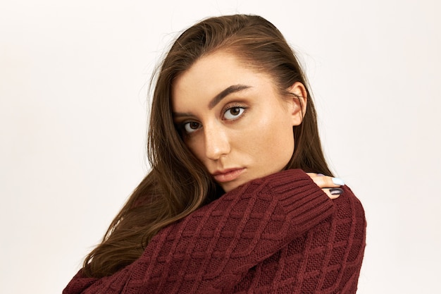 Portrait of attractive cute girl with long dark hair and thoughtful brown eyes having sad look, spending time all alone, hugging herself, dressed in warm knitted sweater. People and lifestyle