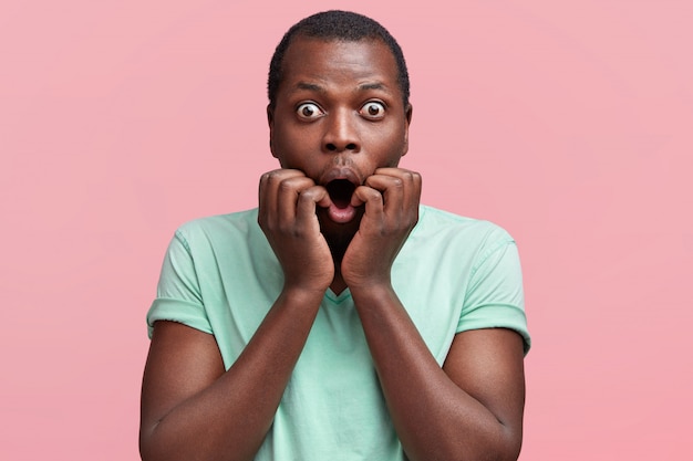 Portrait of attractive dark skinned male looks wih eyes popped out, keeps mouth widely opened, dressed in casual t shirt, poses against pink