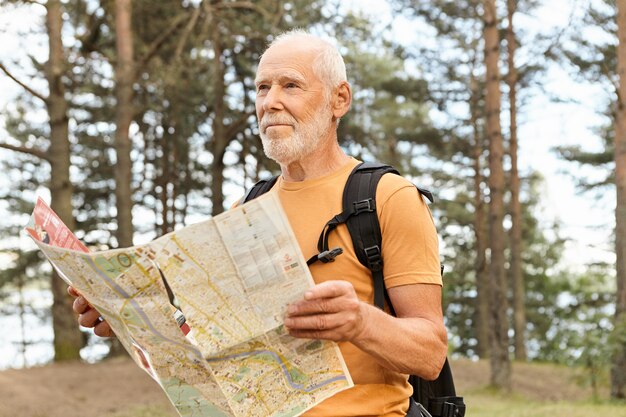 Portrait of attractive elderly male traveler backpacking using map, searching for right route. Bearded senior Caucasian male carrying rucksack thinking where to go, being at crossroads in forest