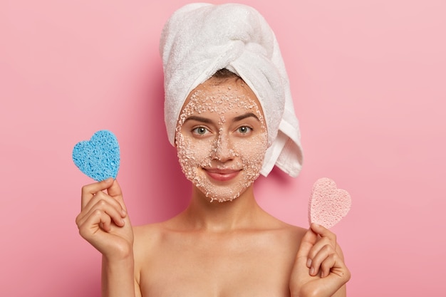 Portrait of attractive European charming woman with sea salt scrub on complexion, wears wrapped soft towel on head, has healthy skin, poses shirtless against rosy background, has tender look