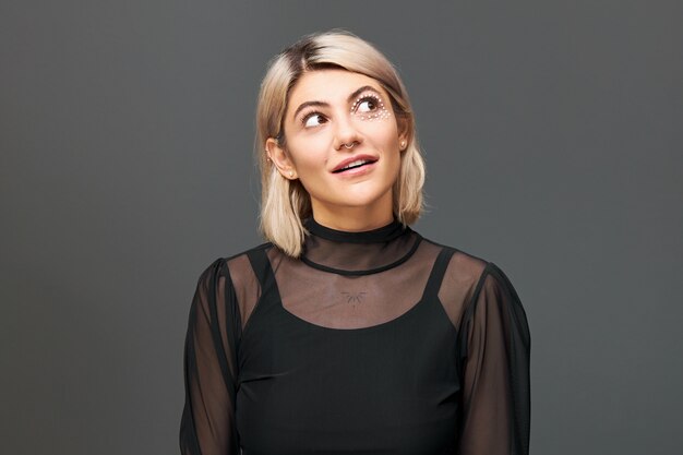 Portrait of attractive fashionable young blonde female dressed in stylish clothes posing isolated looking up with eyes expressing interest and curiosity, opening mouth. Human facial expressions