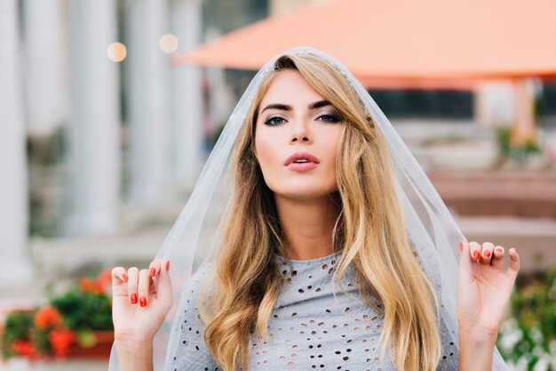 Portrait attractive girl with long blonde hair on street. She holds blue veil covering her head, looking to camera.