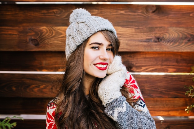 Портрет привлекательной девушки с длинными волосами и красными губами в вязаной шапке на деревянном. Она касается лица рукой в перчатках и улыбается.
