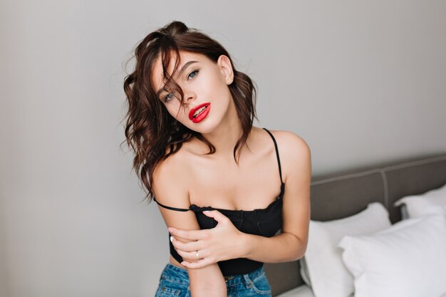 Portrait attractive girl with naked shoulders chilling in modern apartment. Fashionable brunette young model, red lips, relaxing time, fashion.
