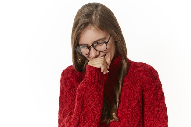 Portrait of attractive joyful girl with loose hair holding hand at her mouth, laughing at joke or funny story. Cute timid young female in eyeglasses and jumper smiling shyly and looking down