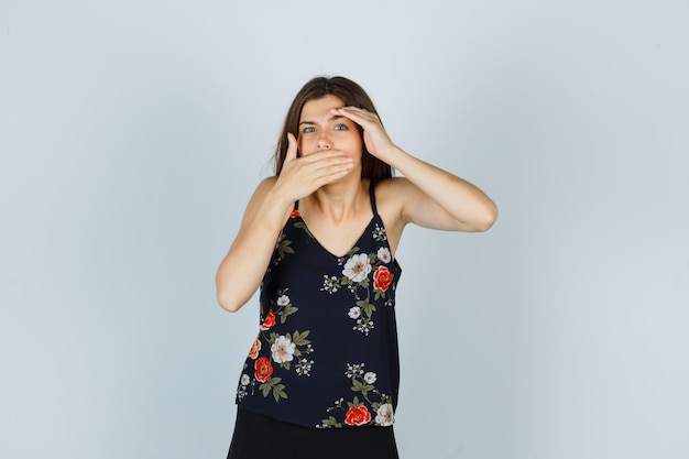 Portrait of attractive lady holding one hand over head, covering mouth with the other hand in blouse and looking troubled front view