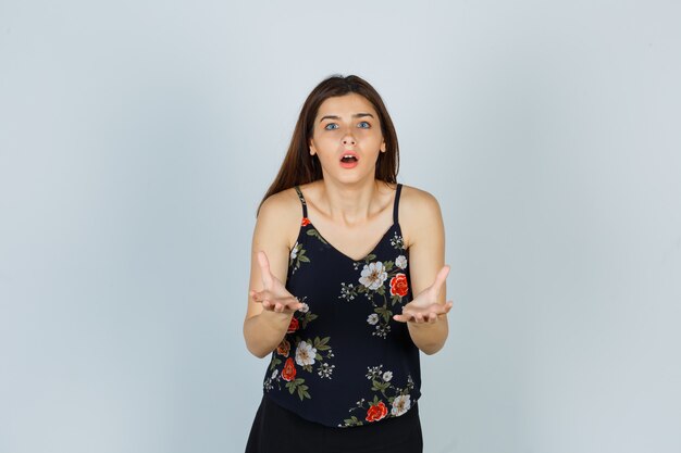 Portrait of attractive lady making asking question gesture in blouse and looking shocked front view