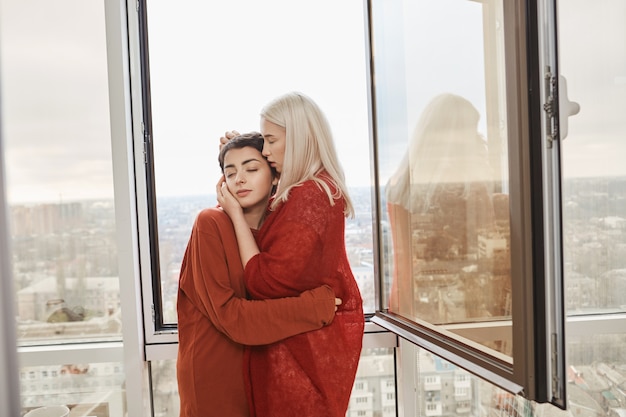 バルコニーに立っている間開いた窓の近くを抱いて、赤い服を着て恋に魅力的なレズビアンのカップルの肖像画。仕事に行く前に一緒に朝を過ごす2人のゲイの女性