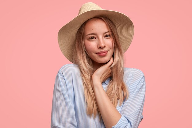 Portrait of attractive lovely young woman wears elegant hat and shirt, holds hand under chin, shows her natural beauty, stands against pink wall